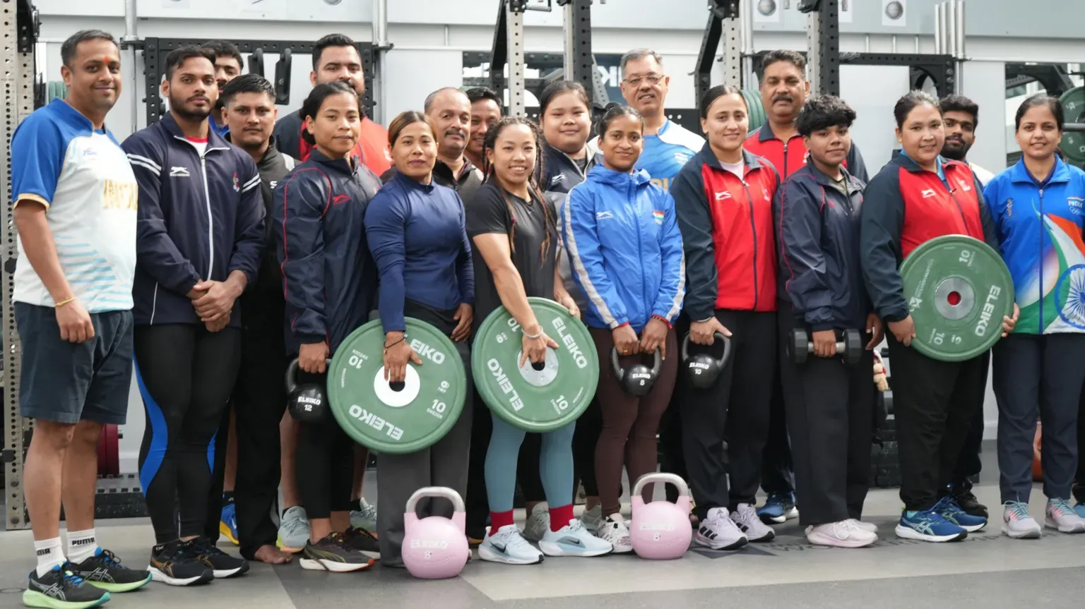 University of Wolverhampton The men and women of the Indian weightlifting team lined up in a gym to pose for a photo, They are wearing gym clothes and some are holding large, round weights.